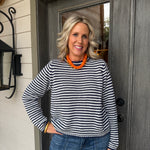 A smiling woman with blonde hair stands in front of a dark gray wooden door, wearing a black and white horizontally striped long-sleeve knit sweater, blue jeans, and a chunky orange beaded necklace.