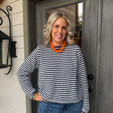 A smiling woman with blonde hair stands in front of a dark gray wooden door, wearing a black and white horizontally striped long-sleeve knit sweater, blue jeans, and a chunky orange beaded necklace.