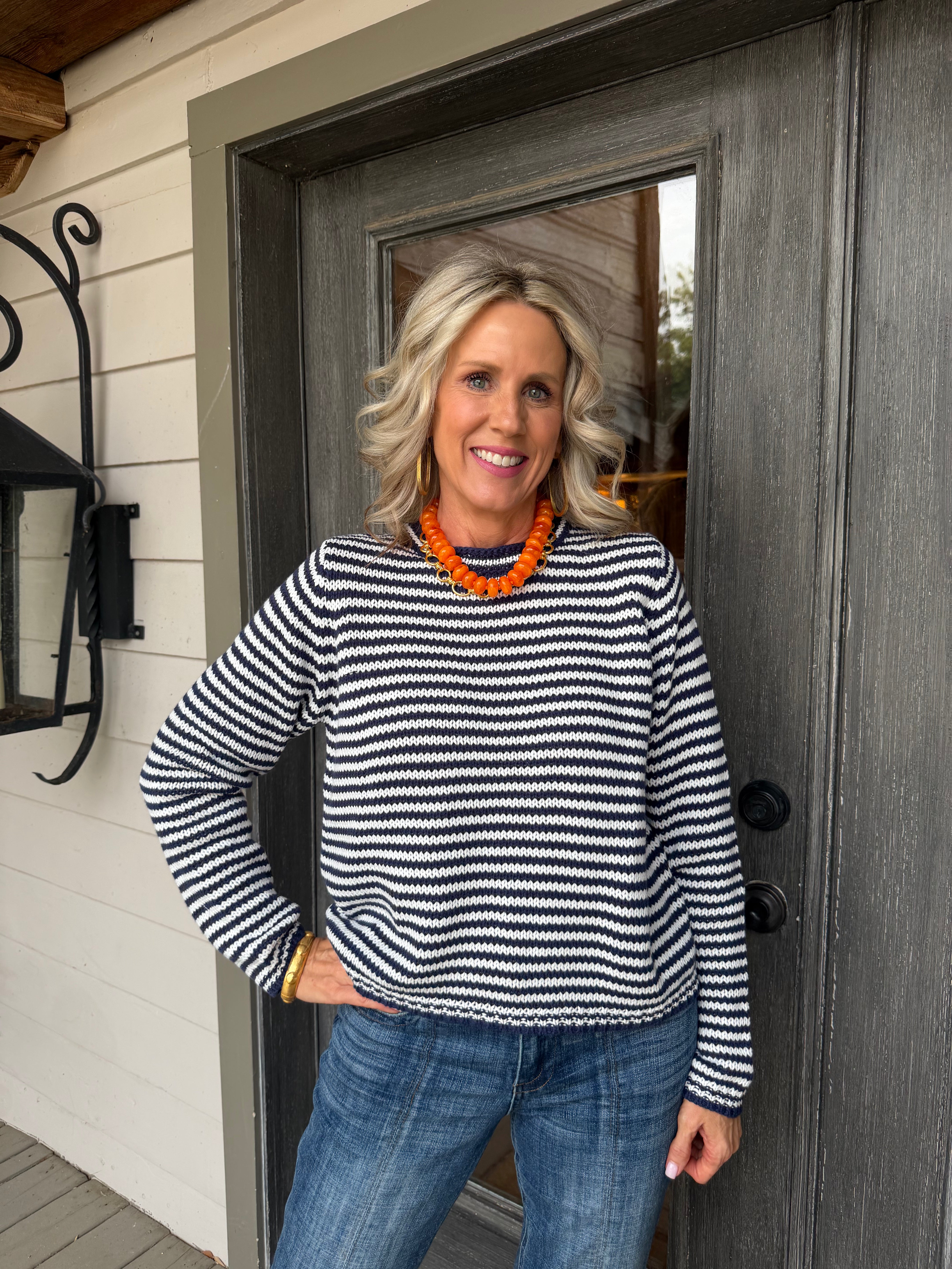 A smiling woman with blonde hair stands in front of a dark gray wooden door, wearing a black and white horizontally striped long-sleeve knit sweater, blue jeans, and a chunky orange beaded necklace.
