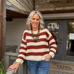 A smiling blonde woman stands on a wooden porch wearing a cream and rust red striped knit sweater, blue jeans, and a chunky statement necklace. A doormat reading 'The Pearl Home Craft' is visible in the background.