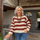 A smiling blonde woman stands on a wooden porch wearing a cream and rust red striped knit sweater, blue jeans, and a chunky statement necklace. A doormat reading 'The Pearl Home Craft' is visible in the background.
