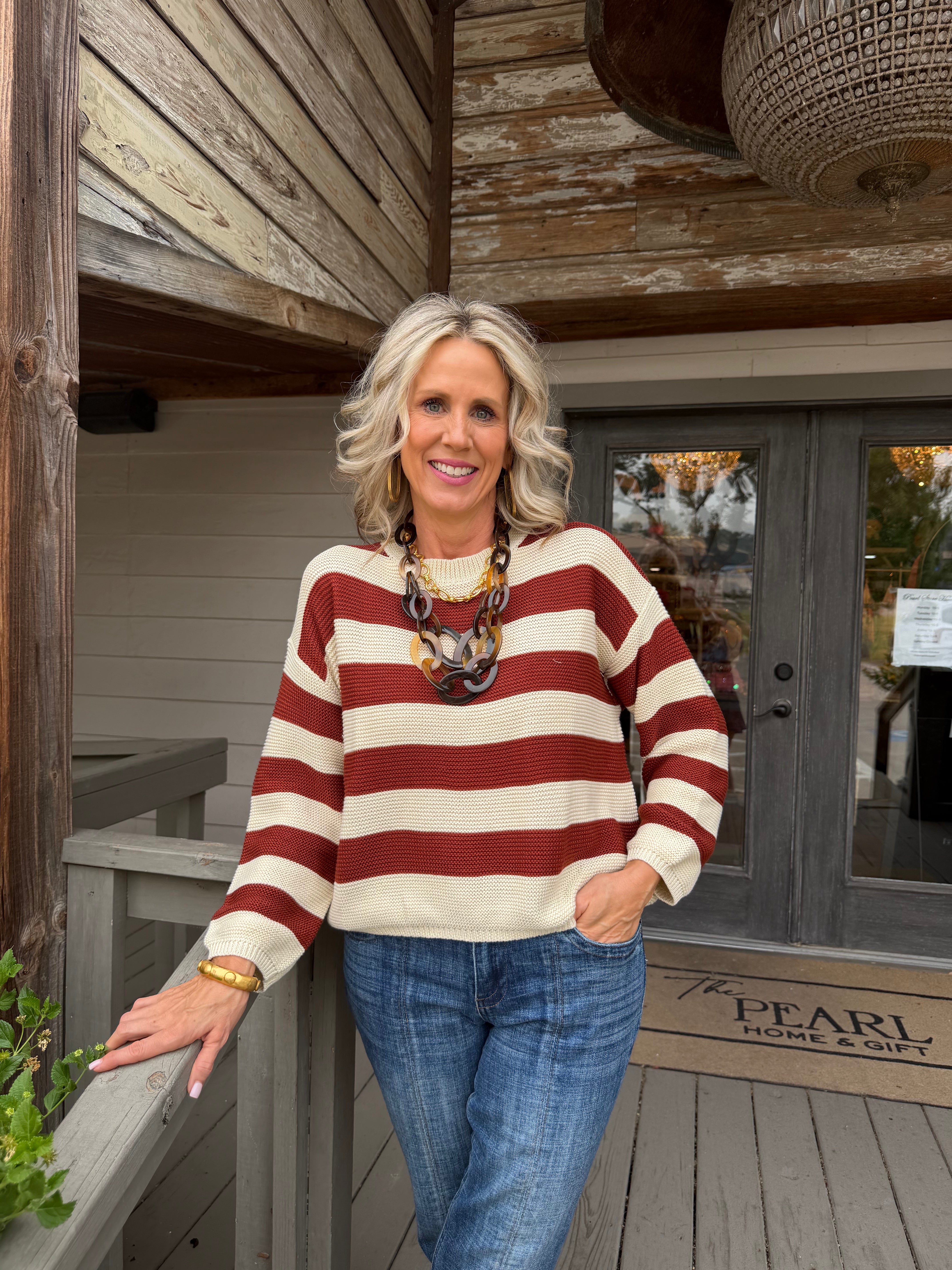 A smiling blonde woman stands on a wooden porch wearing a cream and rust red striped knit sweater, blue jeans, and a chunky statement necklace. A doormat reading 'The Pearl Home Craft' is visible in the background.