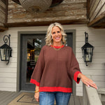 A woman stands on a wooden porch wearing a brown poncho-style sweater with red trim on the cuffs and hem, paired with blue jeans.
