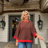 A woman stands on a wooden porch wearing a brown poncho-style sweater with red trim on the cuffs and hem, paired with blue jeans.