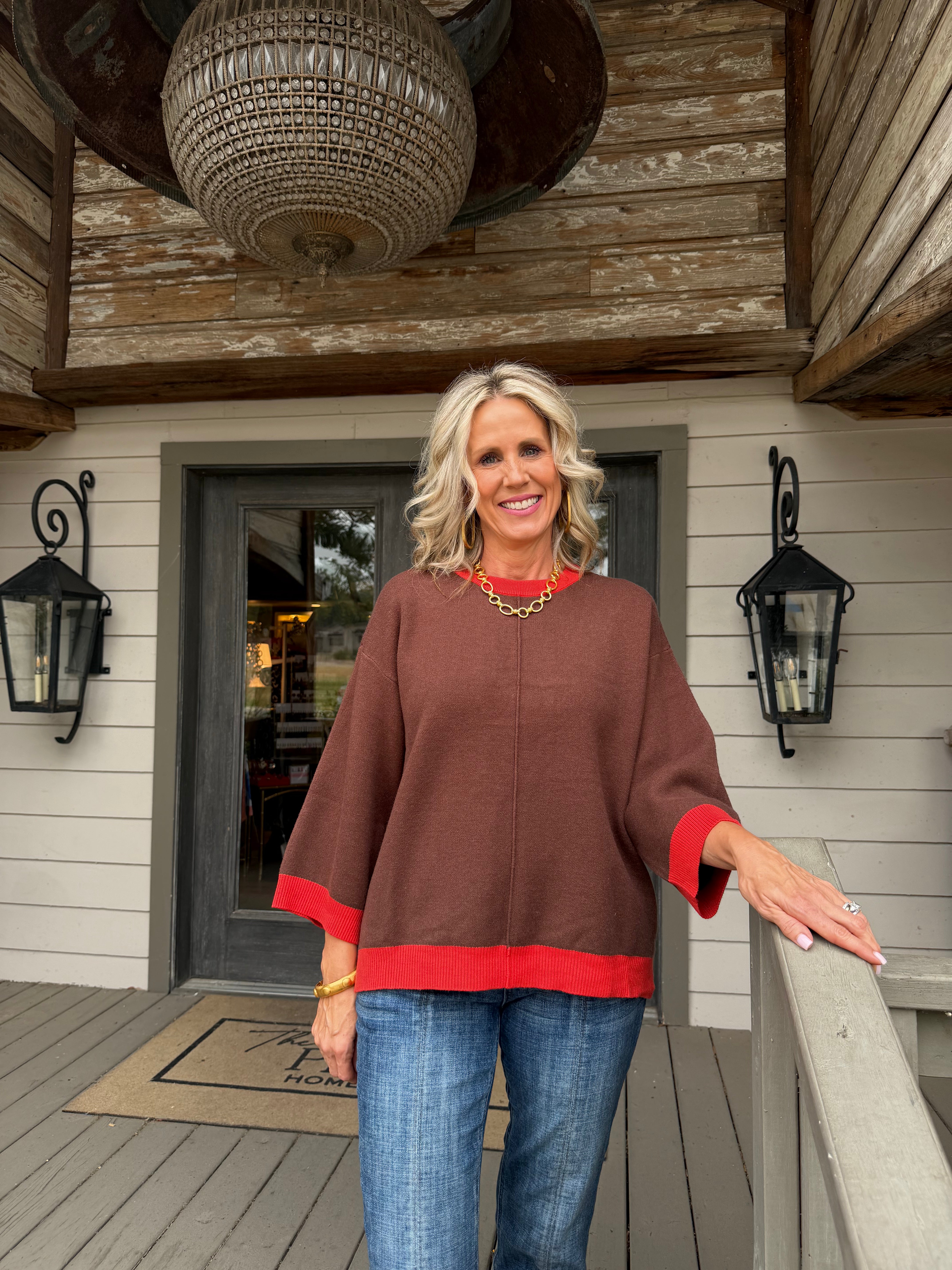 A woman stands on a wooden porch wearing a brown poncho-style sweater with red trim on the cuffs and hem, paired with blue jeans.