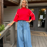 A woman stands on a wooden porch wearing a bright red mock-neck top with white sleeve stripes and wide-leg light blue jeans.