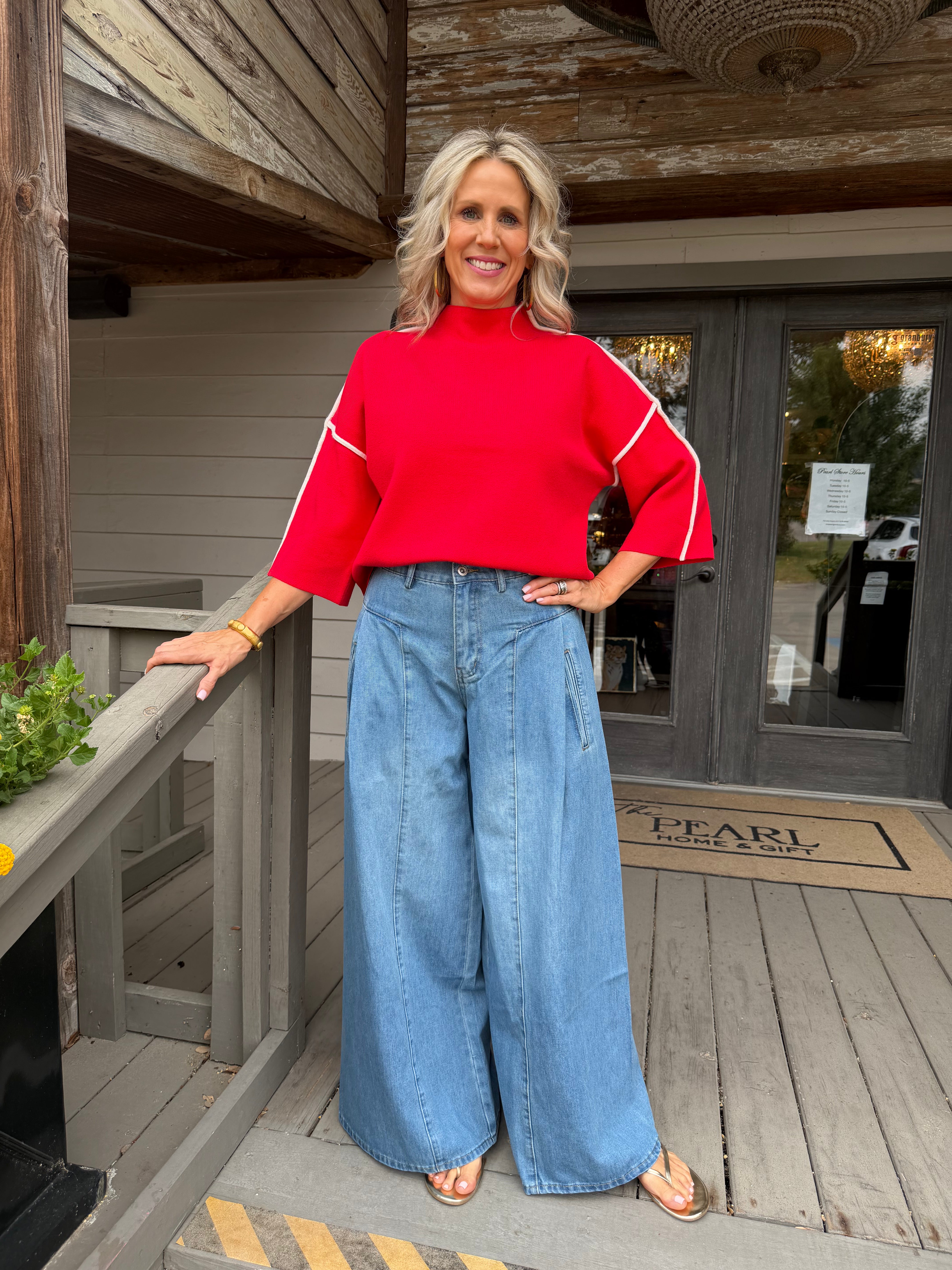 A woman stands on a wooden porch wearing a bright red mock-neck top with white sleeve stripes and wide-leg light blue jeans.