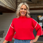 A smiling blonde woman wearing a bright red mock-neck top with white contrast piping on the sleeves, paired with blue jeans, standing outdoors in front of a rustic wooden building.