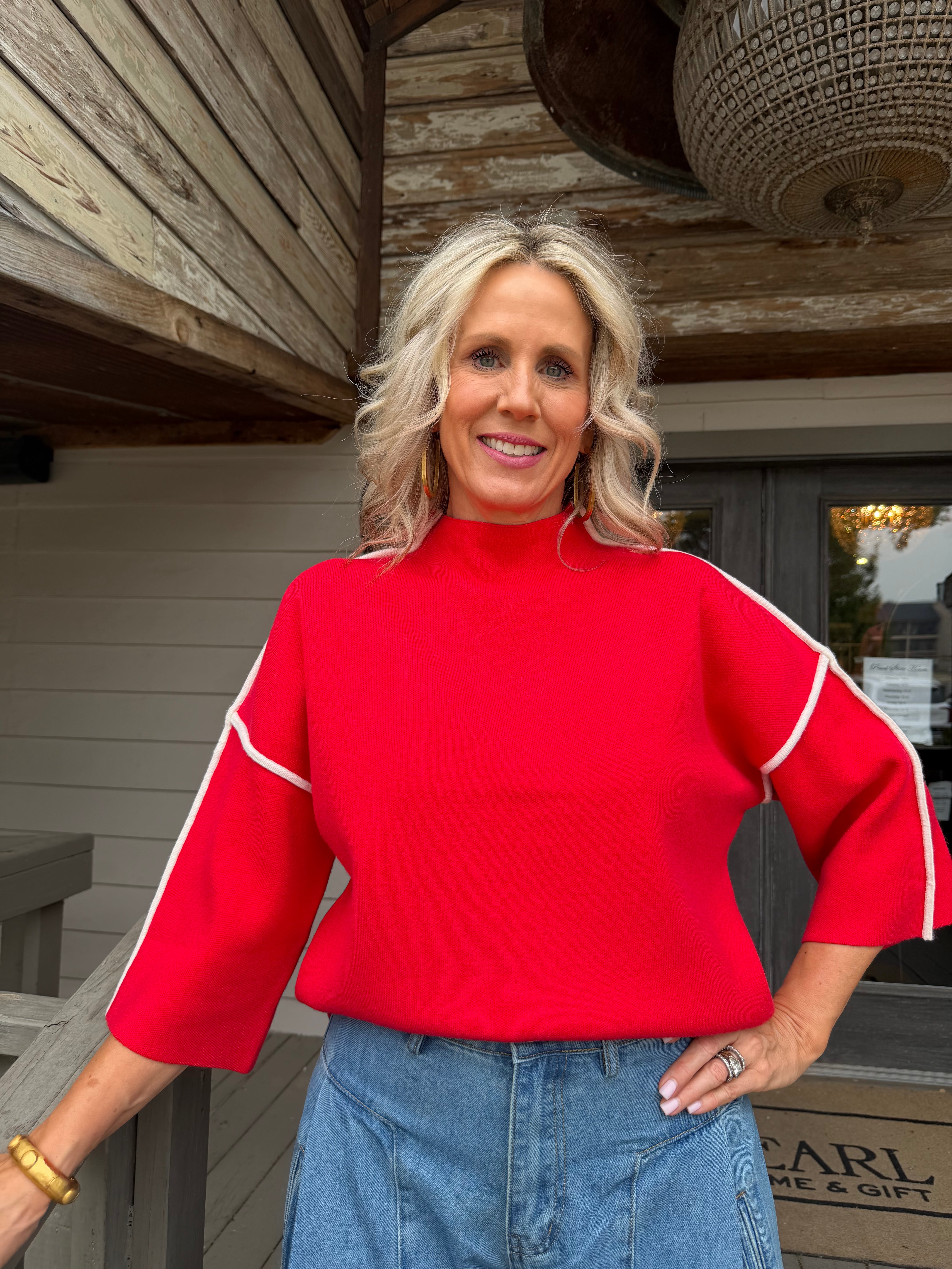 A smiling blonde woman wearing a bright red mock-neck top with white contrast piping on the sleeves, paired with blue jeans, standing outdoors in front of a rustic wooden building.