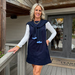 A woman stands on a wooden porch wearing a navy sleeveless dress with a tie-neck detail over a white long-sleeve top. The background shows weathered wood siding, a glass door, and a porch railing.