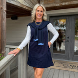 A woman stands on a wooden porch wearing a navy sleeveless dress with a tie-neck detail over a white long-sleeve top. The background shows weathered wood siding, a glass door, and a porch railing.