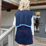 A woman viewed from behind wearing a navy blue sleeveless linen vest with a light blue hem over a white long-sleeve top and navy skirt, standing on a wooden porch.