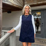 A woman stands on a wooden porch wearing a sleeveless navy blue shift dress over a white long-sleeve top. The porch has light gray wood railings and siding, with a glass door behind her.