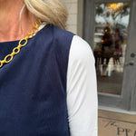 A close-up lifestyle shot of a woman wearing a navy blue sleeveless top over a white long-sleeve shirt, accessorized with a chunky gold chain necklace. She is standing outdoors in front of a building with a gray door.