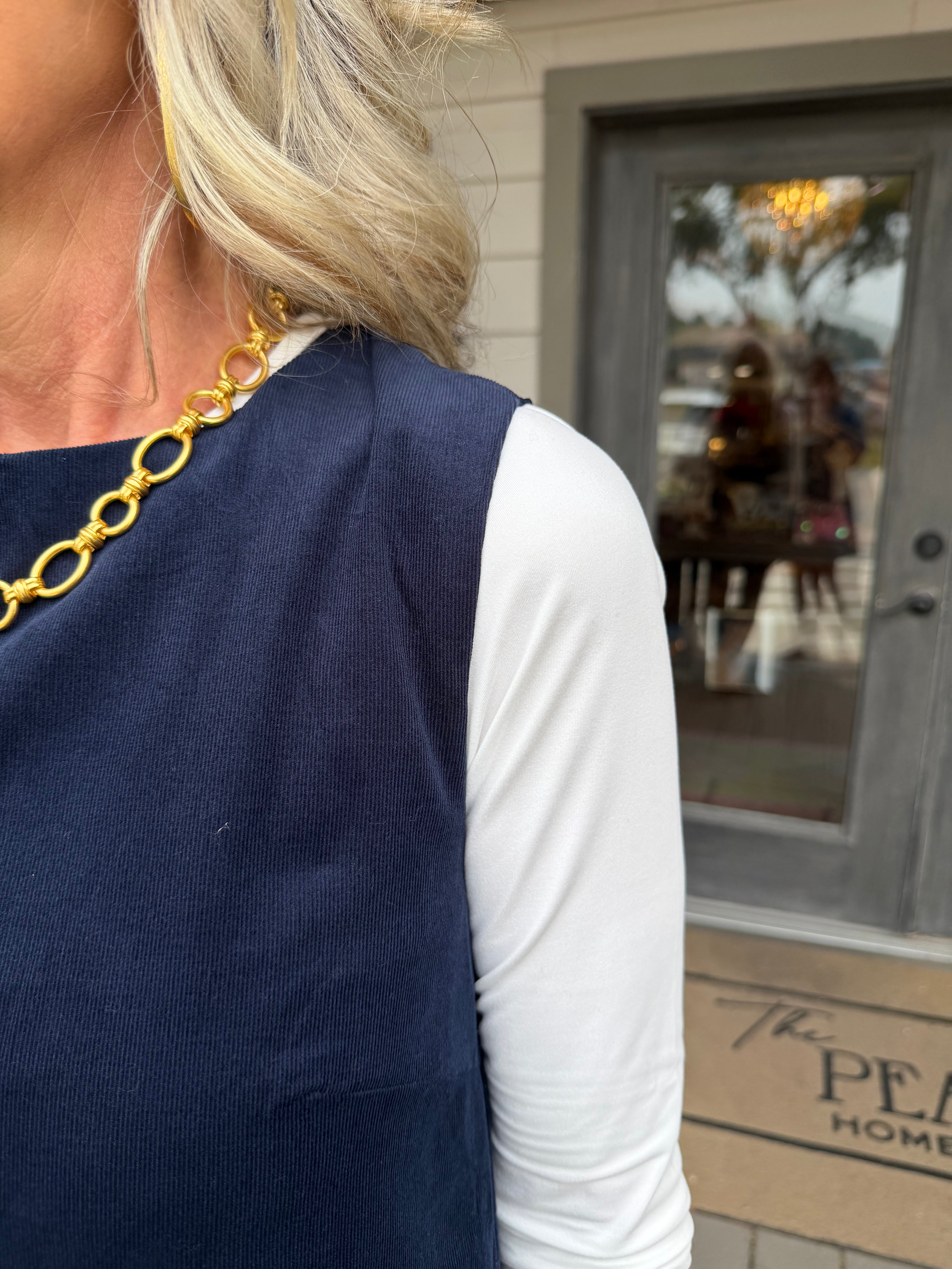 A close-up lifestyle shot of a woman wearing a navy blue sleeveless top over a white long-sleeve shirt, accessorized with a chunky gold chain necklace. She is standing outdoors in front of a building with a gray door.