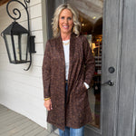 A woman stands on a porch wearing a brown leopard-print cardigan over a white top and blue jeans, positioned in front of a gray door and white siding.