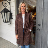 A woman stands on a porch wearing a brown leopard-print cardigan over a white top and blue jeans, positioned in front of a gray door and white siding.