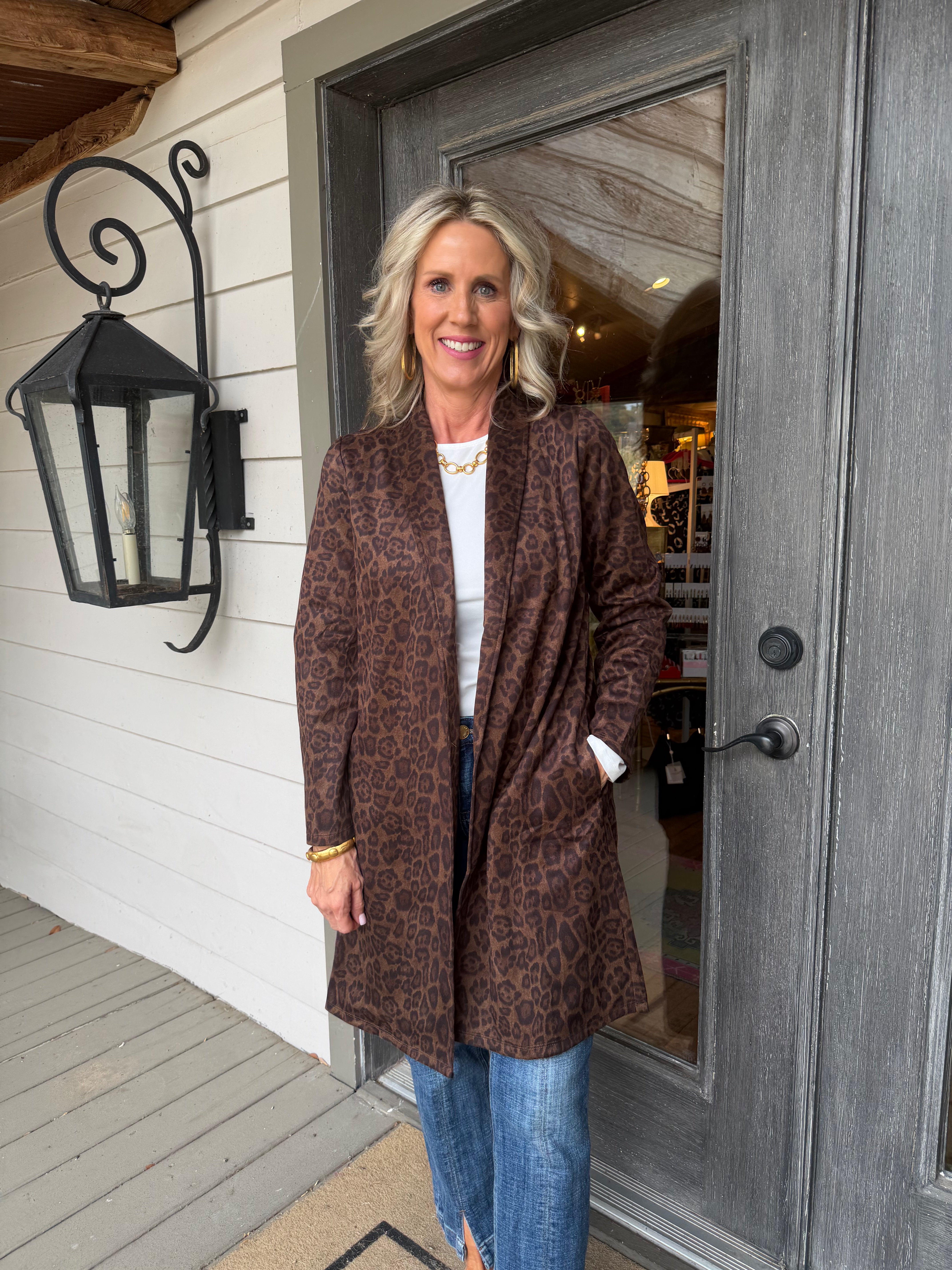 A woman stands on a porch wearing a brown leopard-print cardigan over a white top and blue jeans, positioned in front of a gray door and white siding.