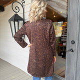 A woman stands on a porch from the back, wearing a brown leopard-print long-sleeve tunic top with blue jeans. The background shows a white wood-paneled wall, a black decorative wall lantern, and a gray wooden door with a glass window.