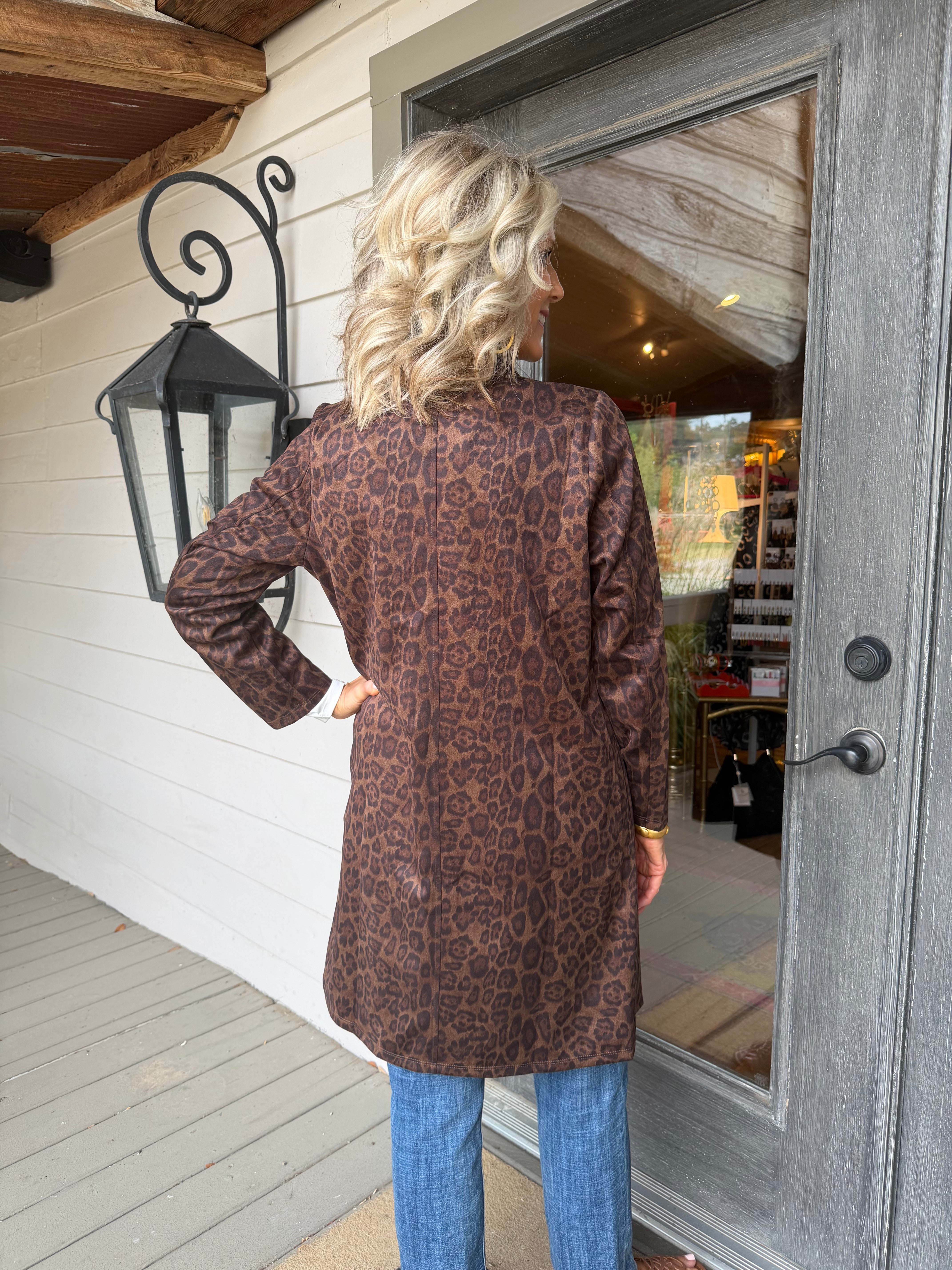 A woman stands on a porch from the back, wearing a brown leopard-print long-sleeve tunic top with blue jeans. The background shows a white wood-paneled wall, a black decorative wall lantern, and a gray wooden door with a glass window.