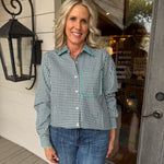A woman with blonde hair stands on a porch wearing a green and white gingham long-sleeve button-up shirt with light blue piping details on the collar, cuffs, and chest pocket, paired with blue jeans.