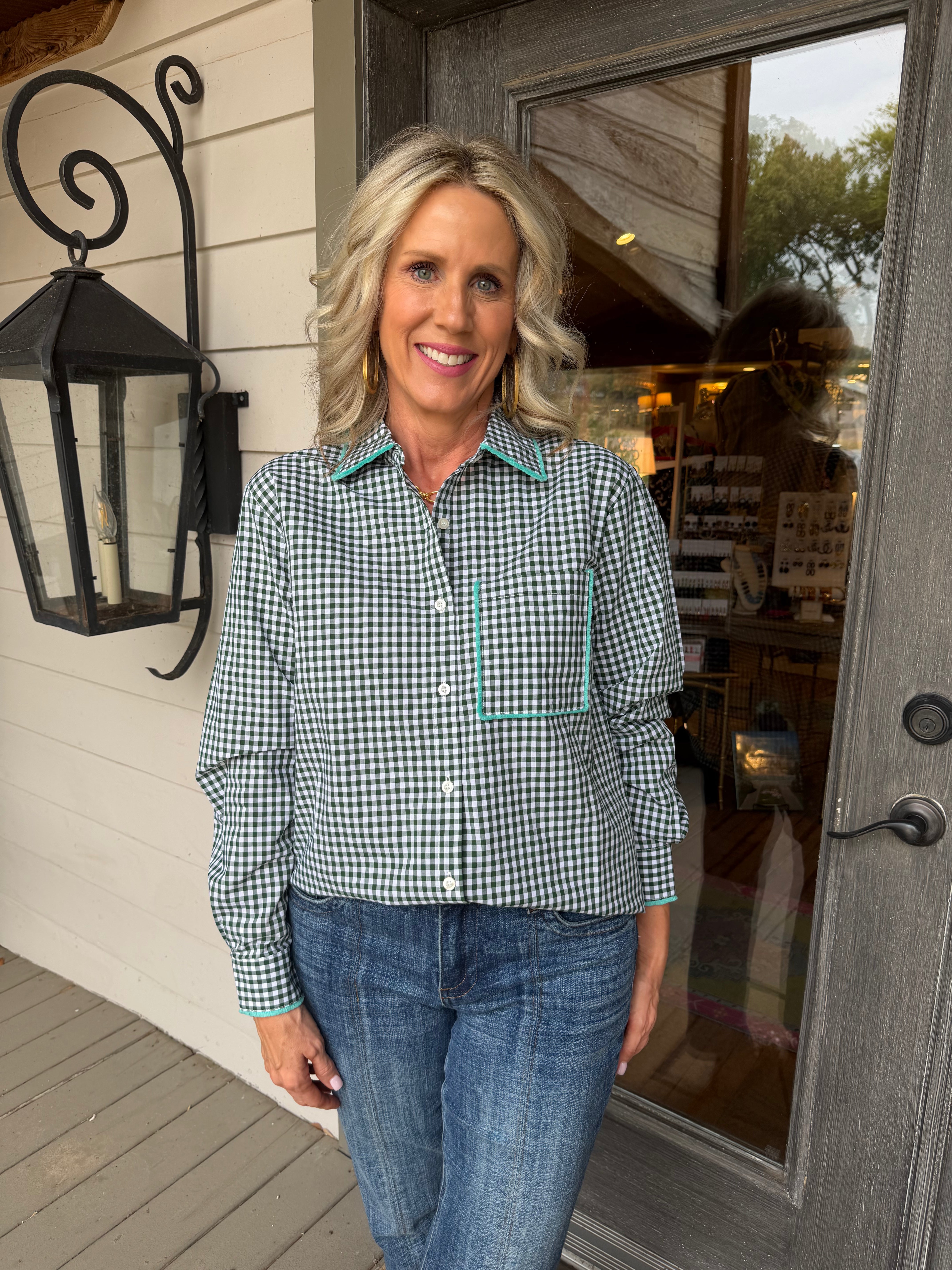 A woman with blonde hair stands on a porch wearing a green and white gingham long-sleeve button-up shirt with light blue piping details on the collar, cuffs, and chest pocket, paired with blue jeans.