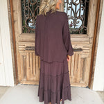 A woman stands with her back to the camera wearing a matching dark plum purple two-piece set consisting of a long-sleeve cropped jacket and a tiered maxi skirt, positioned in front of a rustic wooden door.