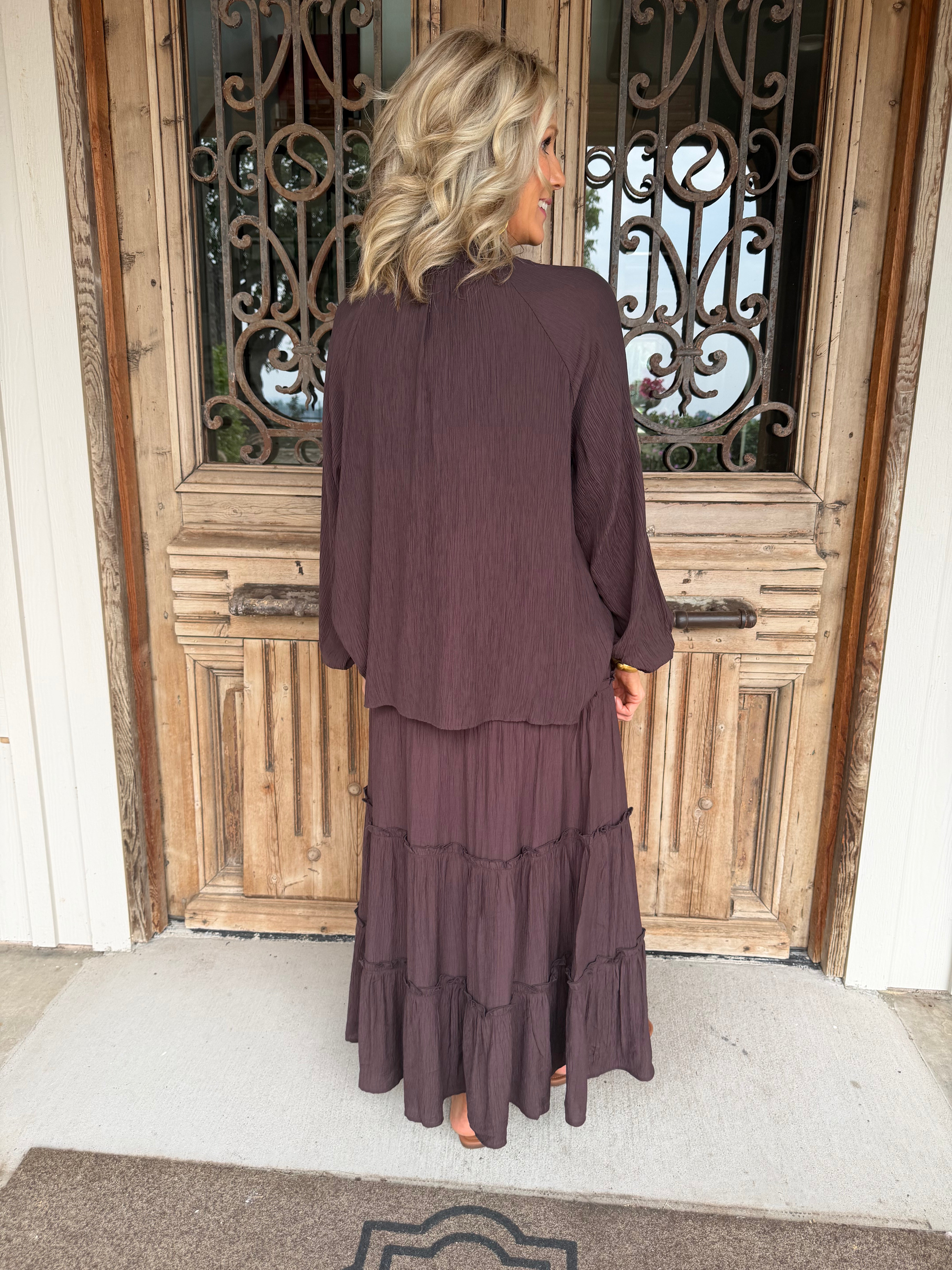 A woman stands with her back to the camera wearing a matching dark plum purple two-piece set consisting of a long-sleeve cropped jacket and a tiered maxi skirt, positioned in front of a rustic wooden door.
