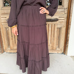 A person wearing a dark brown tiered maxi skirt with a matching long-sleeve button-front top, standing in front of a rustic wooden door.