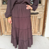 A person wearing a dark brown tiered maxi skirt with a matching long-sleeve button-front top, standing in front of a rustic wooden door.