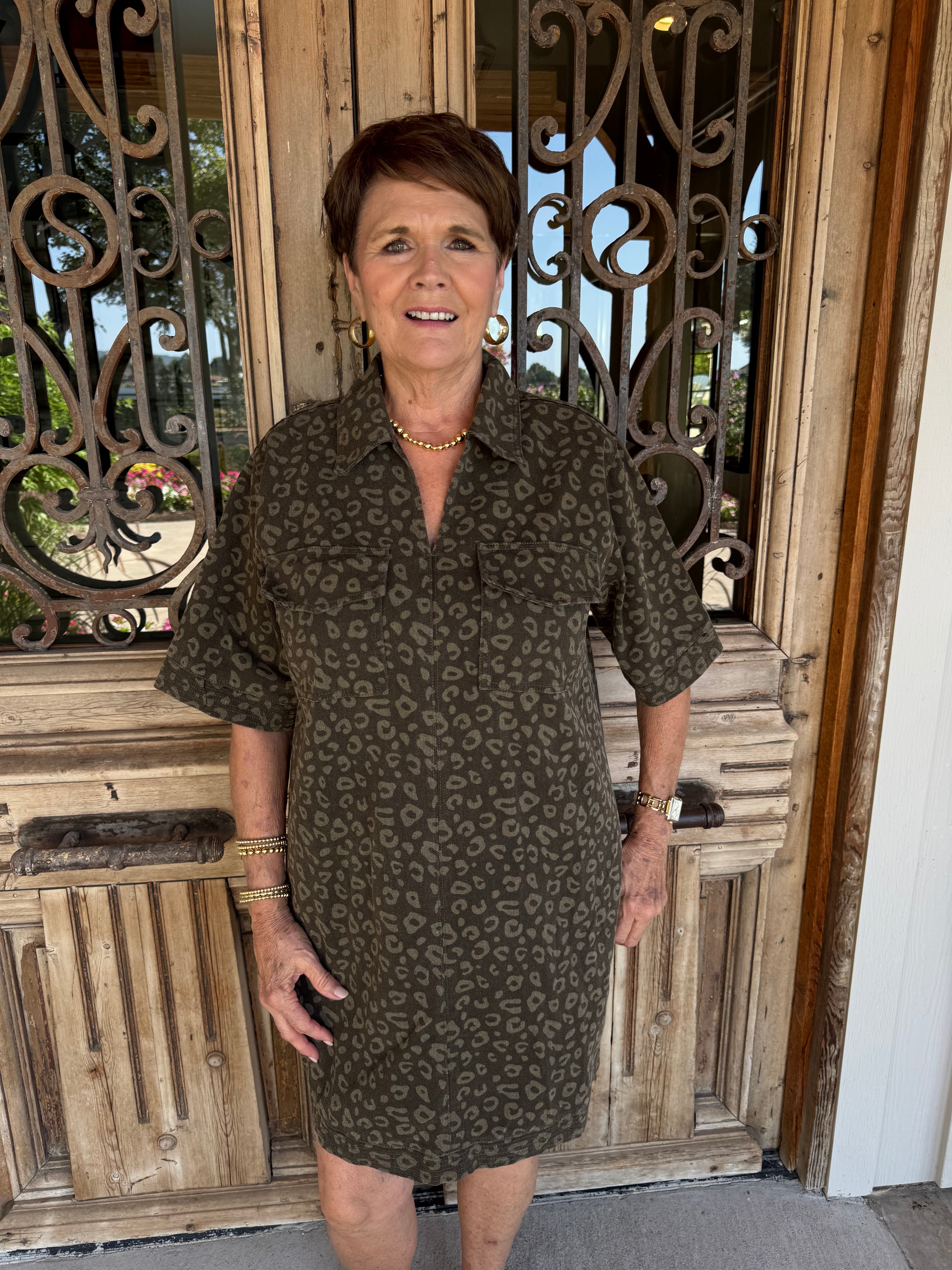 A woman wearing a short-sleeve olive green and brown leopard print dress stands in front of a rustic wooden door with an ornate metal gate.