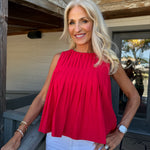 A smiling blonde woman wearing a bright red sleeveless pleated top and white pants, standing on a wooden porch.