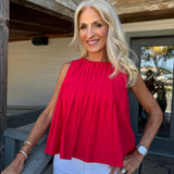 A smiling blonde woman wearing a bright red sleeveless pleated top and white pants, standing on a wooden porch.