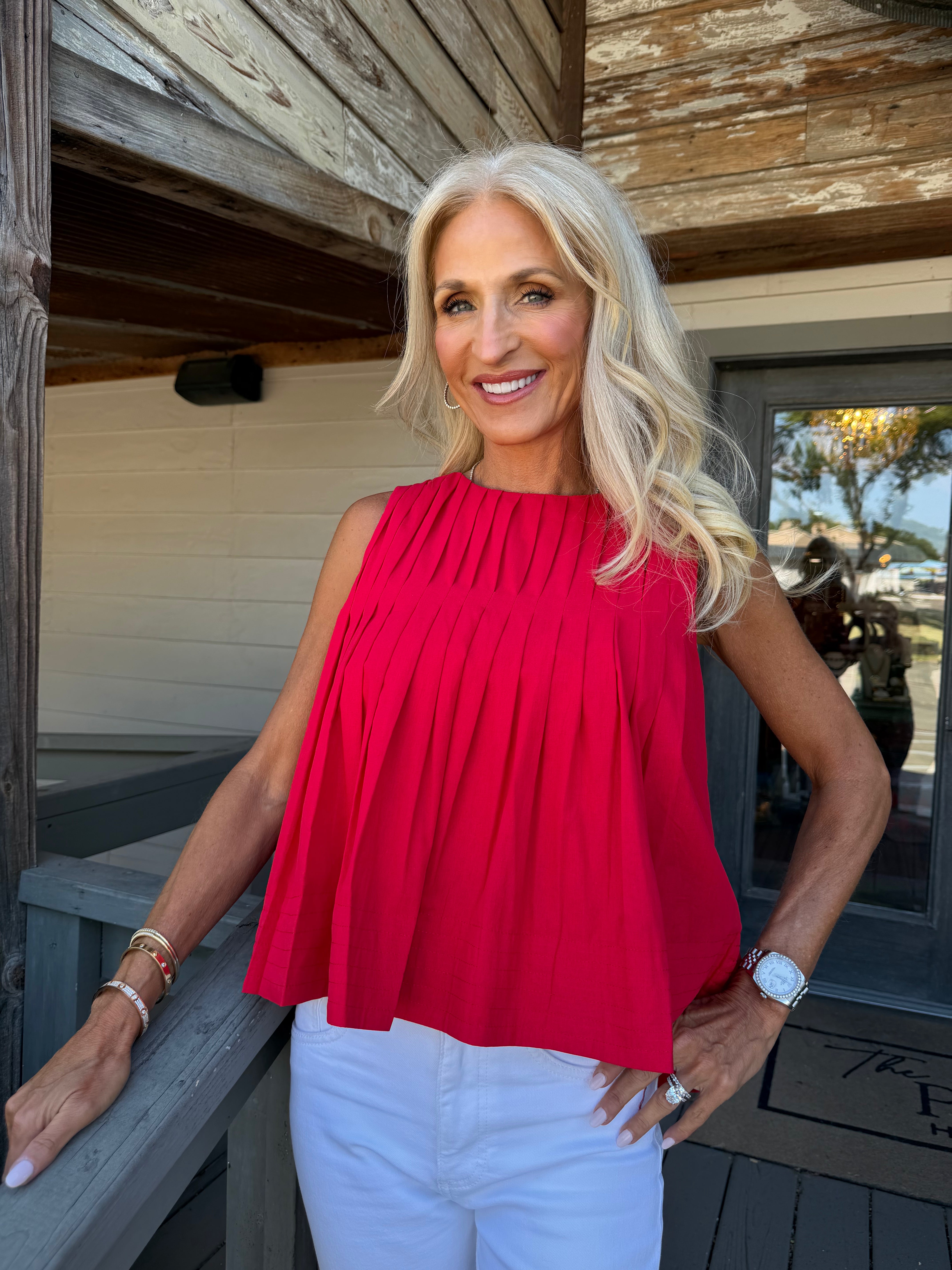 A smiling blonde woman wearing a bright red sleeveless pleated top and white pants, standing on a wooden porch.