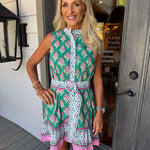 A woman stands on a porch wearing a sleeveless green dress with a pink and white geometric pattern, a high collar, and a matching tie belt.
