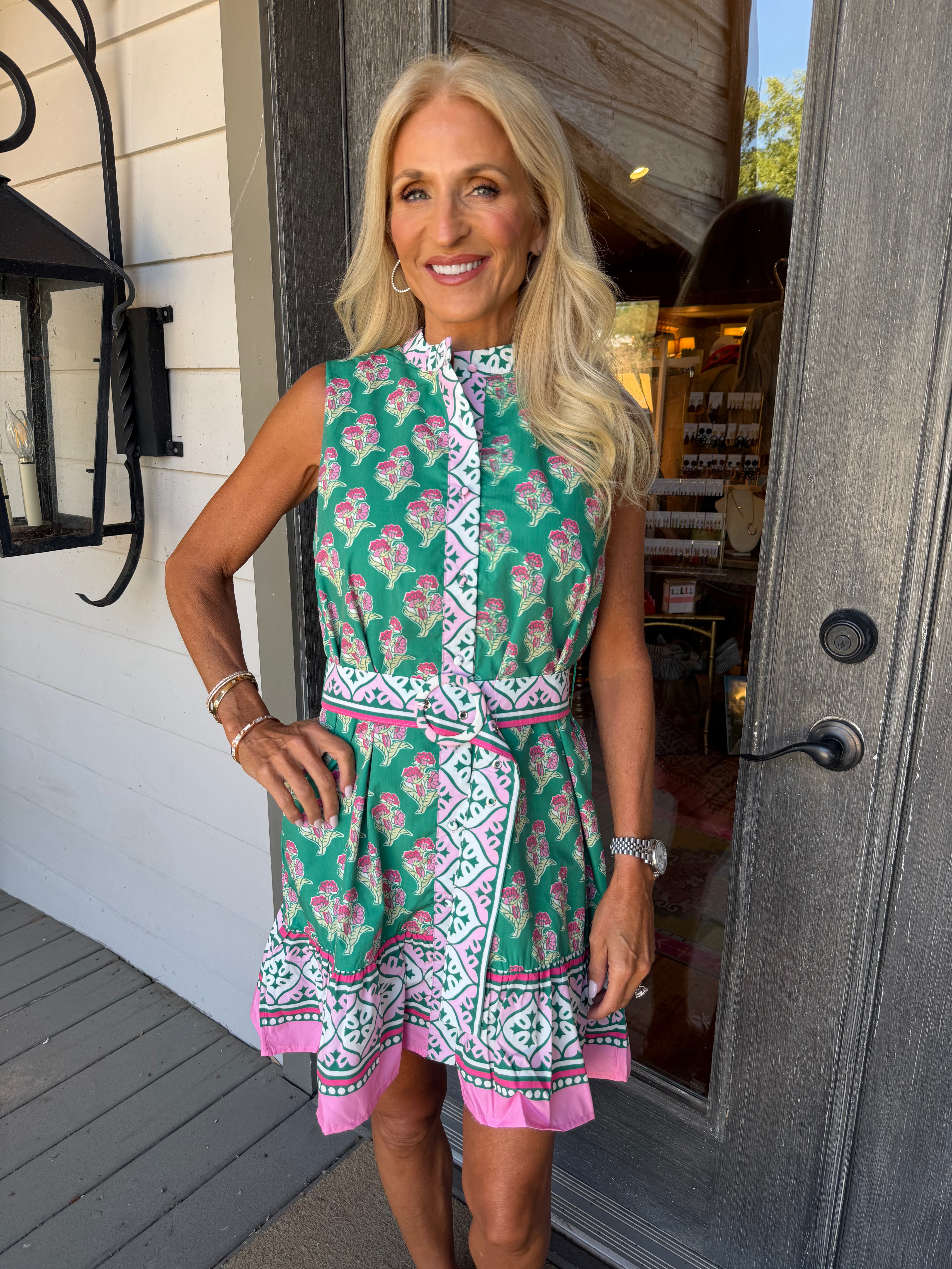 A woman stands on a porch wearing a sleeveless green dress with a pink and white geometric pattern, a high collar, and a matching tie belt.