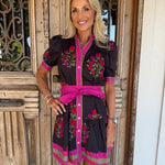 A woman modeling a black short-sleeve dress with bright pink trim, a matching pink waist tie belt, and colorful floral embroidery, standing in front of a rustic wooden door.