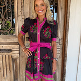 A woman modeling a black short-sleeve dress with bright pink trim, a matching pink waist tie belt, and colorful floral embroidery, standing in front of a rustic wooden door.
