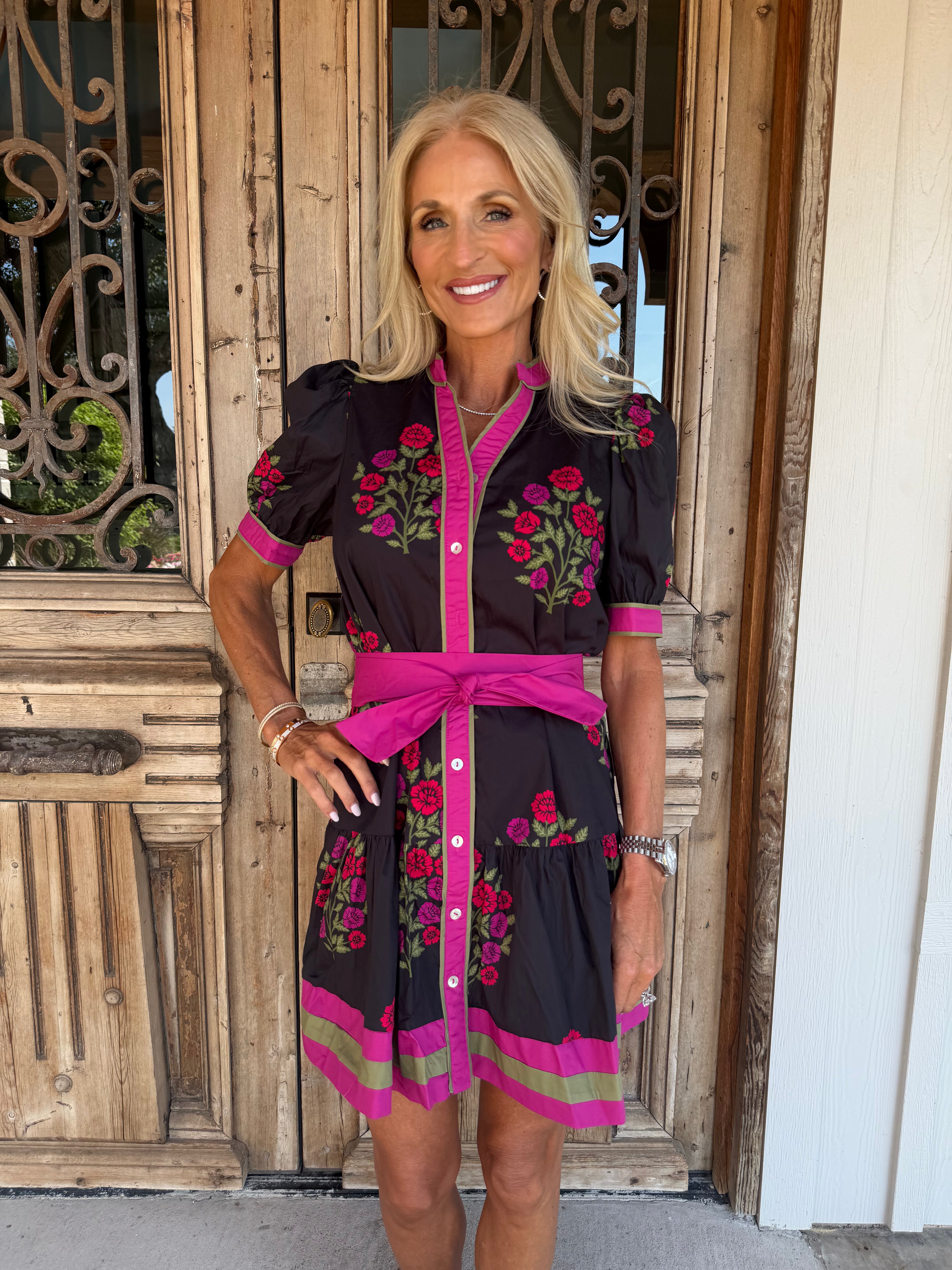 A woman modeling a black short-sleeve dress with bright pink trim, a matching pink waist tie belt, and colorful floral embroidery, standing in front of a rustic wooden door.