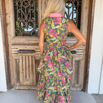 A woman stands with her back to the camera wearing a sleeveless tiered midi dress with a vibrant floral print in pink, yellow, and green on a dark green base, featuring a contrasting light pink collar. She is positioned in front of a rustic wooden door with decorative metal grates.