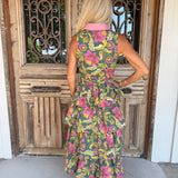 A woman stands with her back to the camera wearing a sleeveless tiered midi dress with a vibrant floral print in pink, yellow, and green on a dark green base, featuring a contrasting light pink collar. She is positioned in front of a rustic wooden door with decorative metal grates.