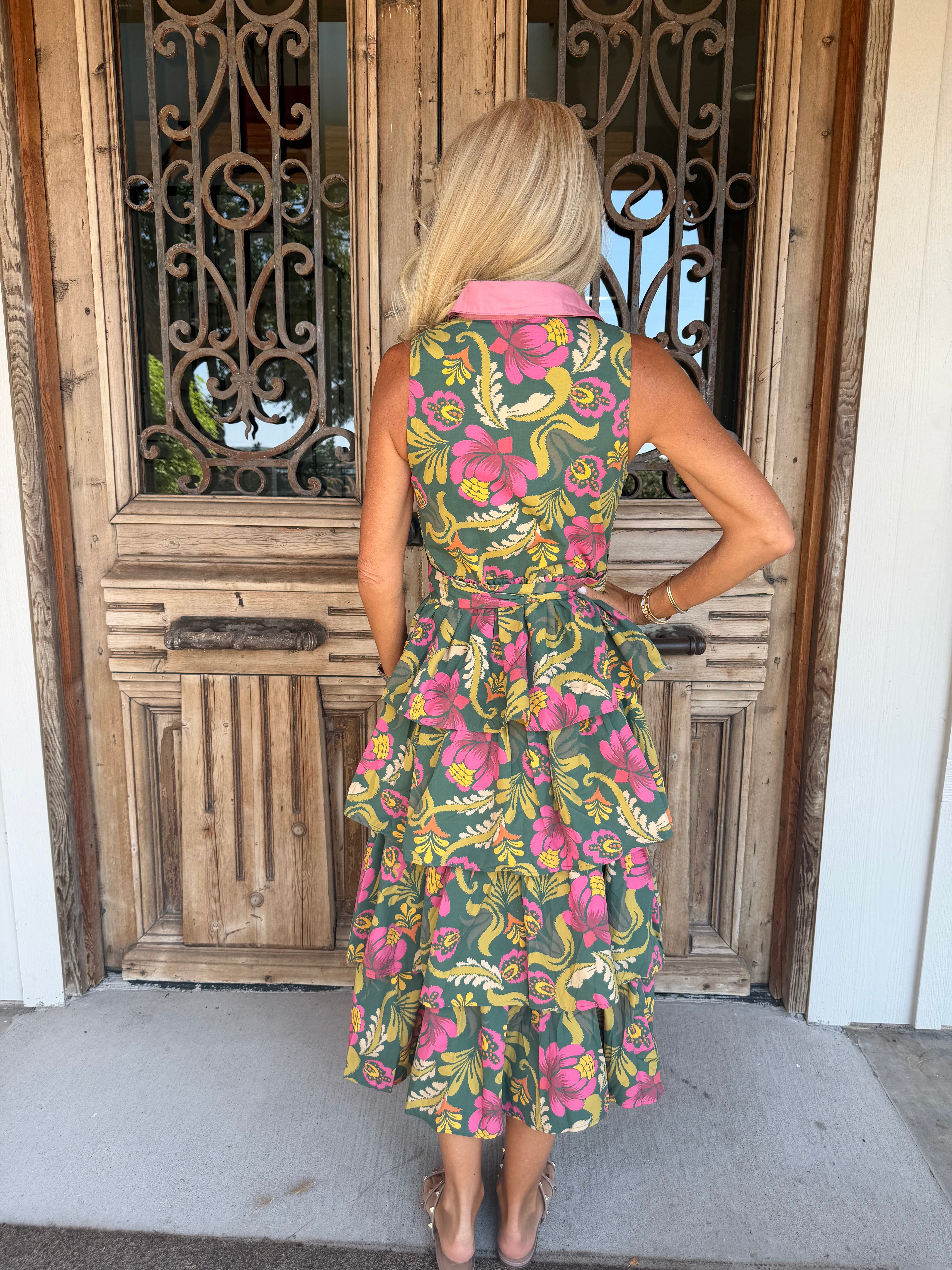 A woman stands with her back to the camera wearing a sleeveless tiered midi dress with a vibrant floral print in pink, yellow, and green on a dark green base, featuring a contrasting light pink collar. She is positioned in front of a rustic wooden door with decorative metal grates.