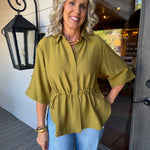 A smiling blonde woman stands on a porch wearing a loose olive green short-sleeve button-up top with a gathered waist and light blue jeans.