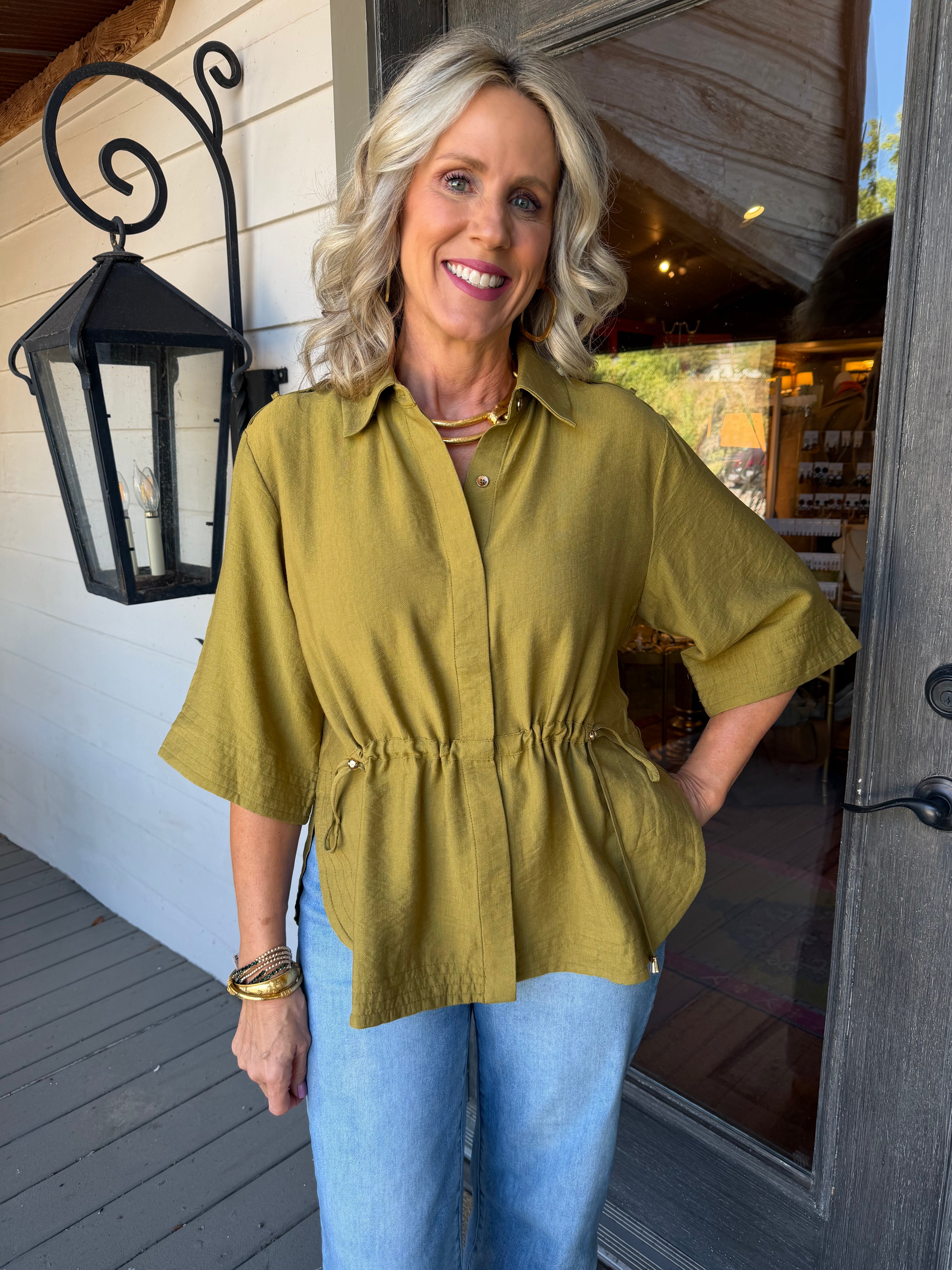 A smiling blonde woman stands on a porch wearing a loose olive green short-sleeve button-up top with a gathered waist and light blue jeans.