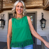 A smiling blonde woman stands on a porch wearing a bright green sleeveless top with fringe trim at the hem, paired with light blue jeans and gold jewelry.