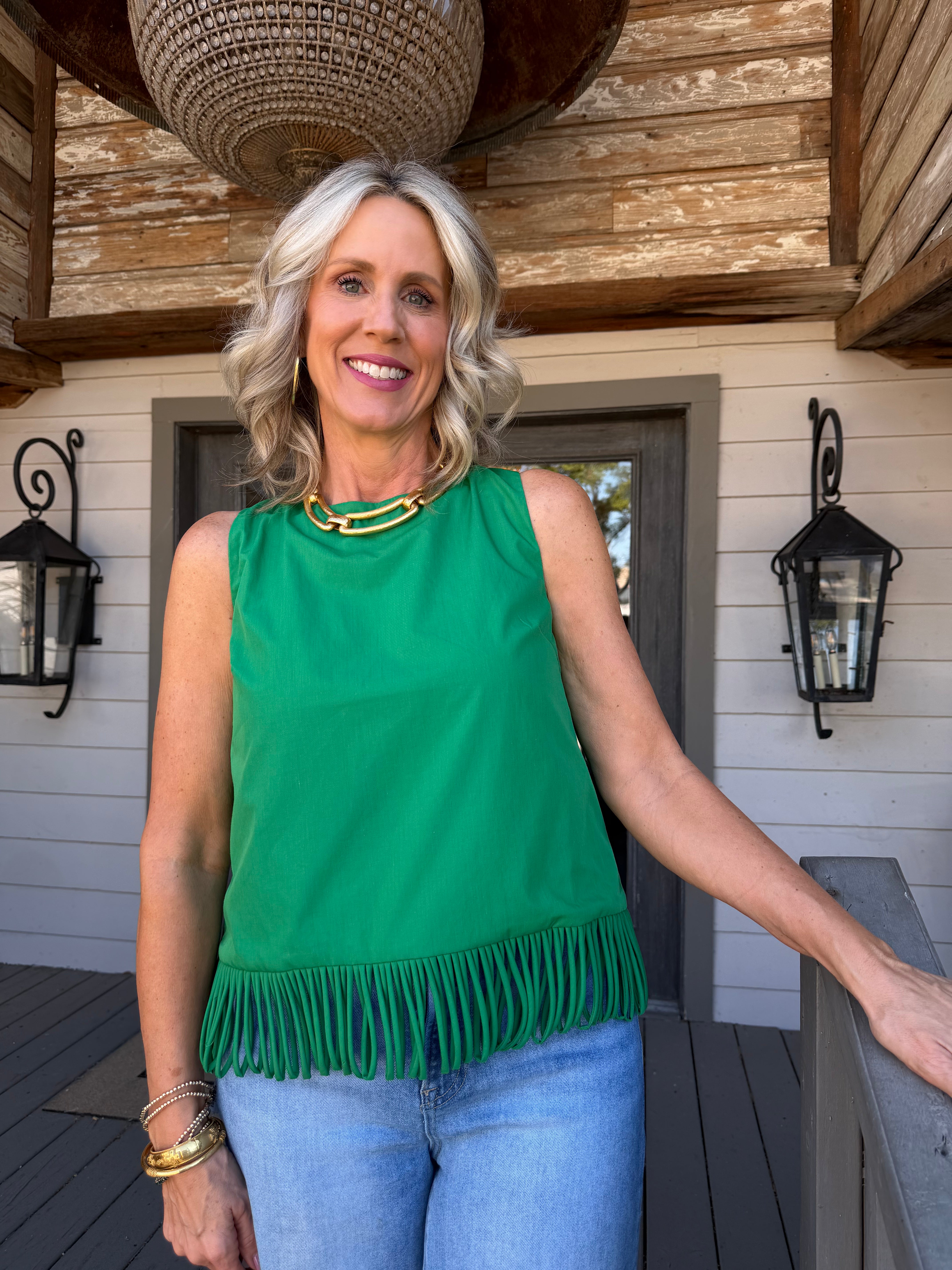 A smiling blonde woman stands on a porch wearing a bright green sleeveless top with fringe trim at the hem, paired with light blue jeans and gold jewelry.