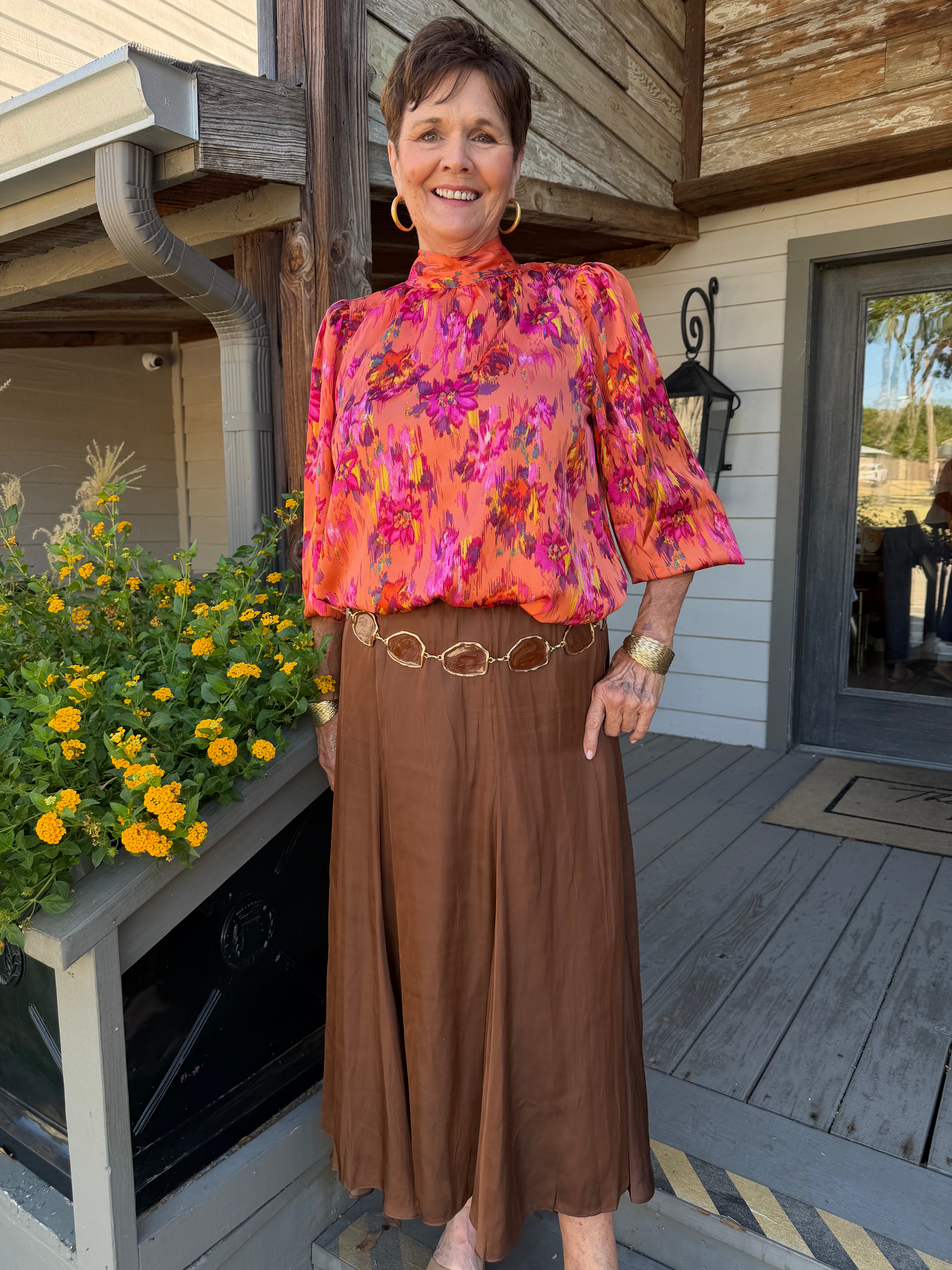 Woman standing outside in front of flowers with orange and pink floral blouse on long sleeves bow details 
