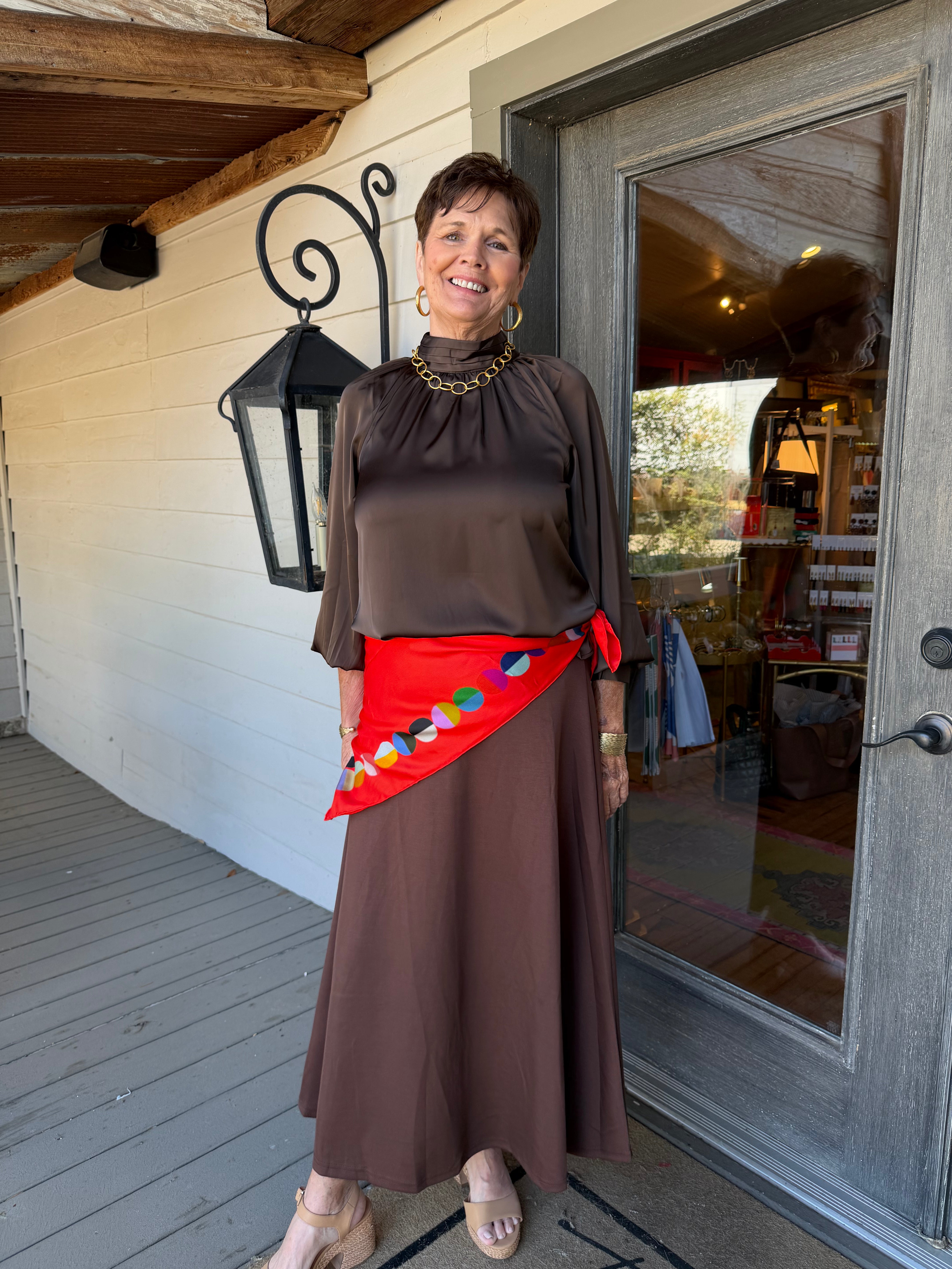 A woman stands on a porch wearing a brown satin-like long-sleeve top and matching maxi skirt, with a bright red wrap-style sash featuring colorful embroidered details tied at the waist.