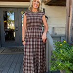 A woman stands on a rustic wooden porch wearing a brown and beige plaid sleeveless maxi dress with a tiered waist. The background features weathered wood siding, a gray door, and a potted green plant with yellow flowers.