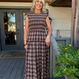 A woman stands on a rustic wooden porch wearing a brown and beige plaid sleeveless maxi dress with a tiered waist. The background features weathered wood siding, a gray door, and a potted green plant with yellow flowers.